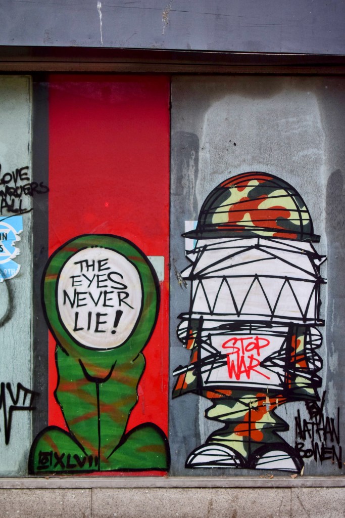 Street art featuring two graffiti characters: one green figure with the text 'THE EYES NEVER LIE!' and another character wearing a camouflage hat with the text 'STOP WAR'.
