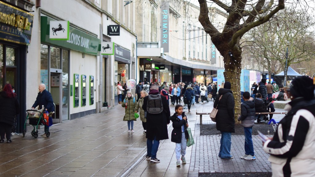A busy city street with a variety of shops including Lloyds Bank and Primark. People are walking along the sidewalk, some with shopping bags, and a tree is visible in the foreground.