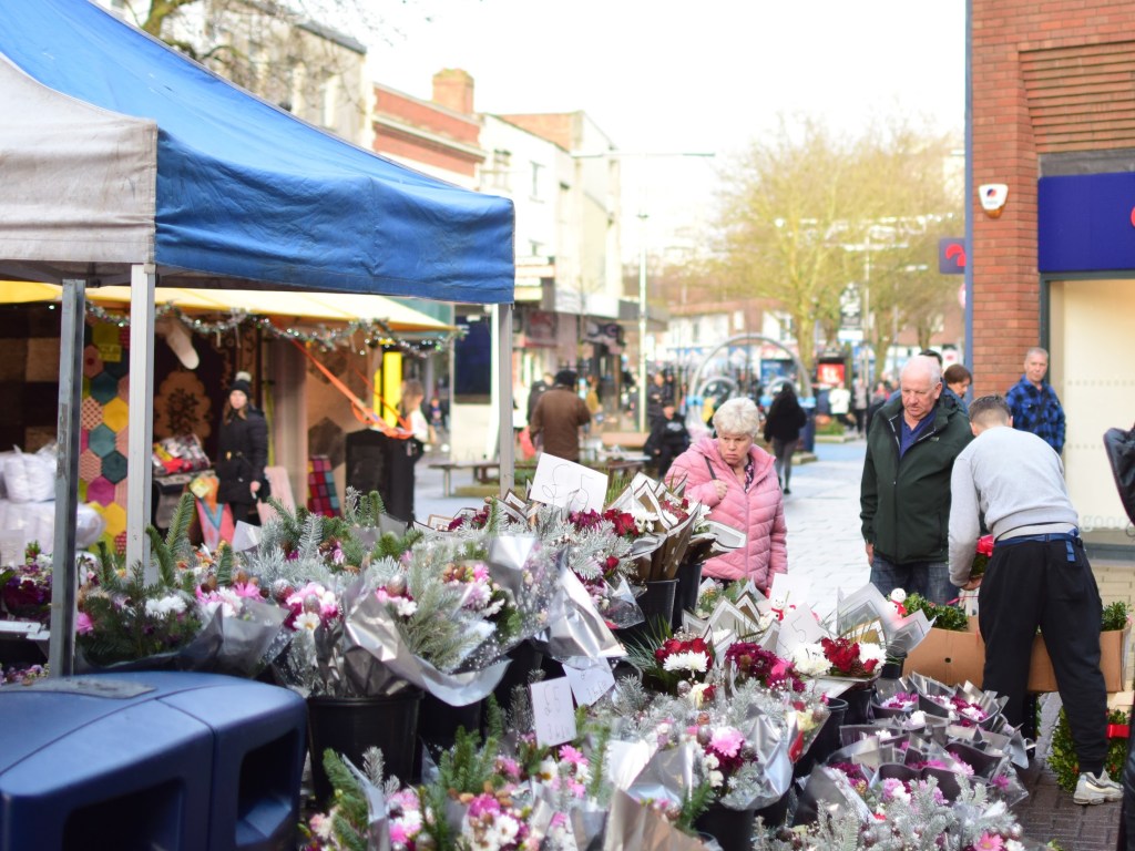 A busy market scene with various flower arrangements for sale, shoppers engaging with vendors, and colorful tents in the background.