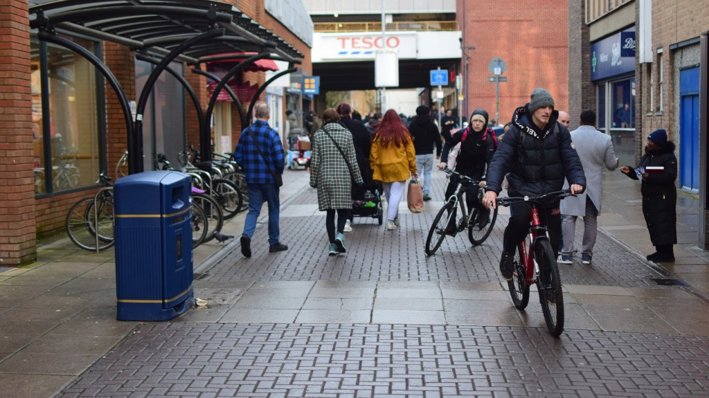 A busy street scene with people walking and cyclists biking in a shopping area. A blue trash can is visible, and a Tesco store is in the background.