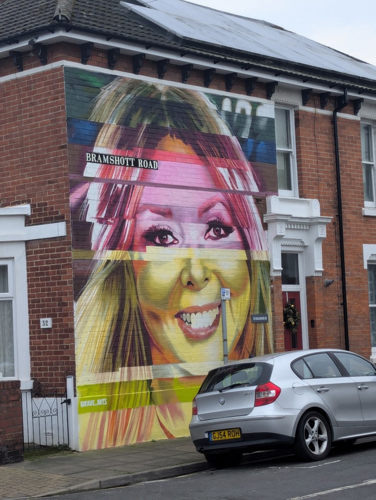 A colorful street mural featuring a smiling woman with long hair, displayed on the side of a brick building, with the text 'BRAMSHOTT ROAD' above her image.